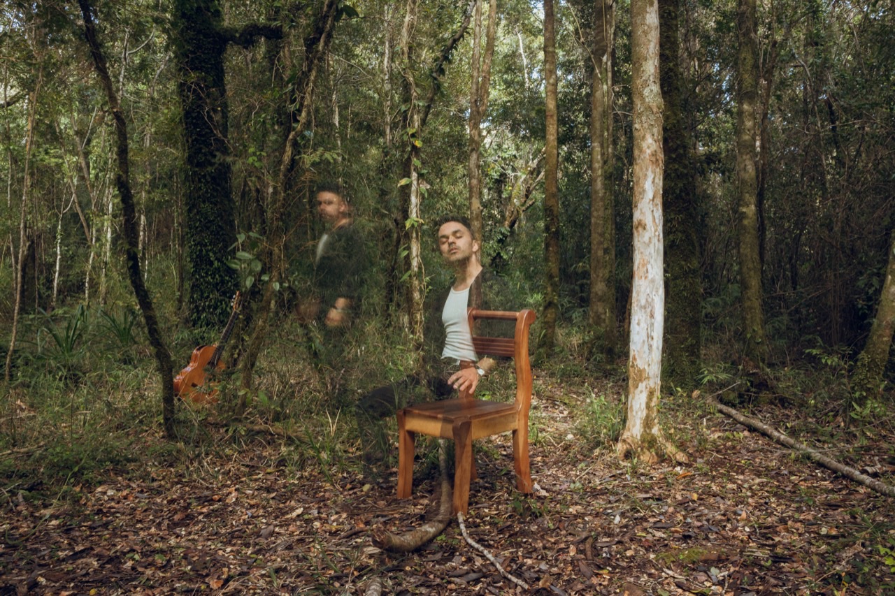 Camilo Eque sentado junto a una silla en el bosque, con una guitarra visible